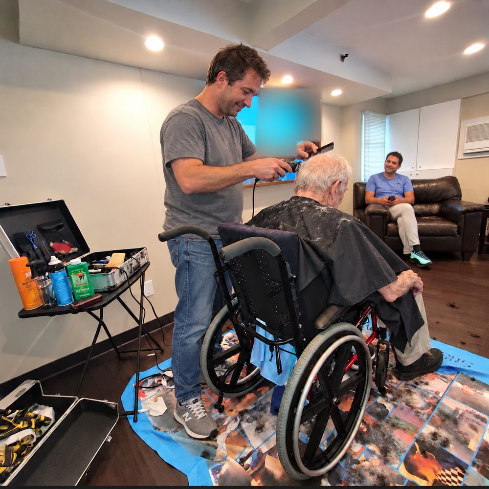A barber cuts an older man's hair while he sits in his wheelchair.